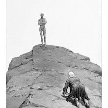 A man climbs a rocky peak on his hands and knees as an almost faceless creature waits at the top