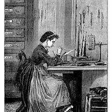 A woman sitting at a work table is blowing a glass imitation pearl in the flame of a gas burner