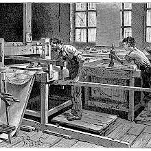 View of a workshop where a man uses a hand block to impress motifs on a strip of wallpaper