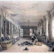 View of the gallery of Naworth Castle, where a man sits reading as a suit of armor lies beside him