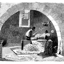 Two workers at a soap factory are seen through an arch crushing blocks of soda with shovels