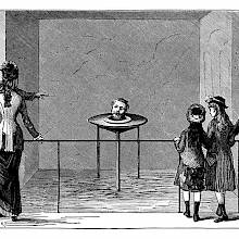 An small audience looks at a table on which the severed head of a man is staring back and talking