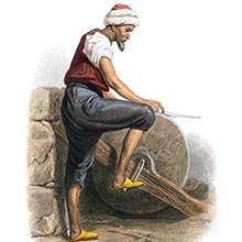 A man is seen from the side sharpening a knife on a grindstone operated by foot