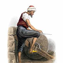 A man is seen from the side sharpening a knife on a grindstone operated by foot