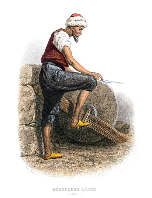 A man is seen from the side sharpening a knife on a grindstone operated by foot