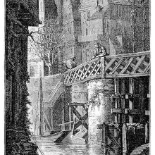 In a city, a woman leans on the railing of a bridge seen from the bank of the river