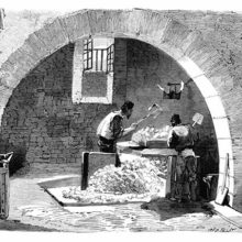 Two workers at a soap factory are seen through an arch crushing blocks of soda with shovels