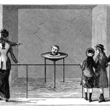 An small audience looks at a table on which the severed head of a man is staring back and talking